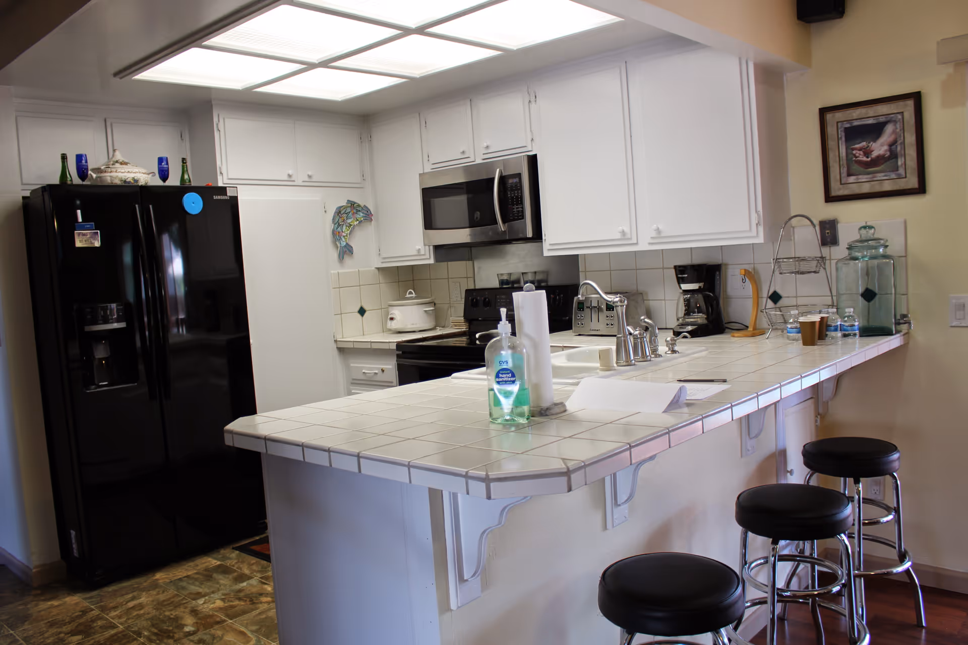 A kitchen with white cabinets and tiled countertops. There is a black refrigerator, a microwave above a stove, a coffee maker, toaster, and a crockpot on the counter. Three black cushioned bar stools are positioned along the counter. Various items including hand sanitizer, paper towels, and cups are on the counter. A framed picture hangs on the wall.