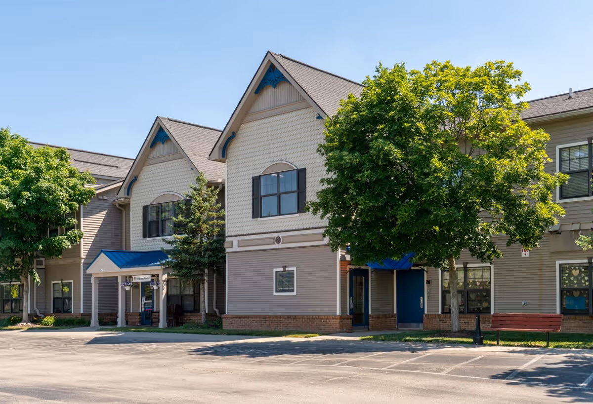 Exterior view of Commonwealth Senior Living at New Baltimore building on a clear sunny day, featuring beige siding with blue accents, multiple windows, a blue awning over the entrance, green trees, and a red bench near the parking area.