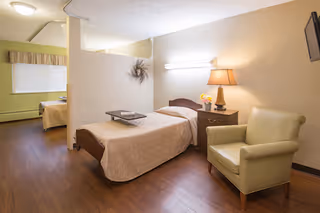 A nursing facility room with a single bed covered in a beige quilt, a nightstand with a lamp and flowers, a comfortable armchair, and a wall-mounted TV. The room has wooden flooring and soft lighting, with a partial view of another bed in an adjacent area separated by a half wall.