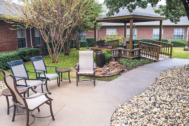Outdoor patio area with several chairs and small tables arranged on a concrete surface. A wooden gazebo with a railing is visible in the background, surrounded by green grass, trees, and shrubs. The building with red brick walls and multiple windows is seen behind the gazebo.