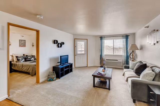 Carpeted living room with a sofa, coffee table, TV on a stand, a window air conditioner, and an open doorway to a bedroom.