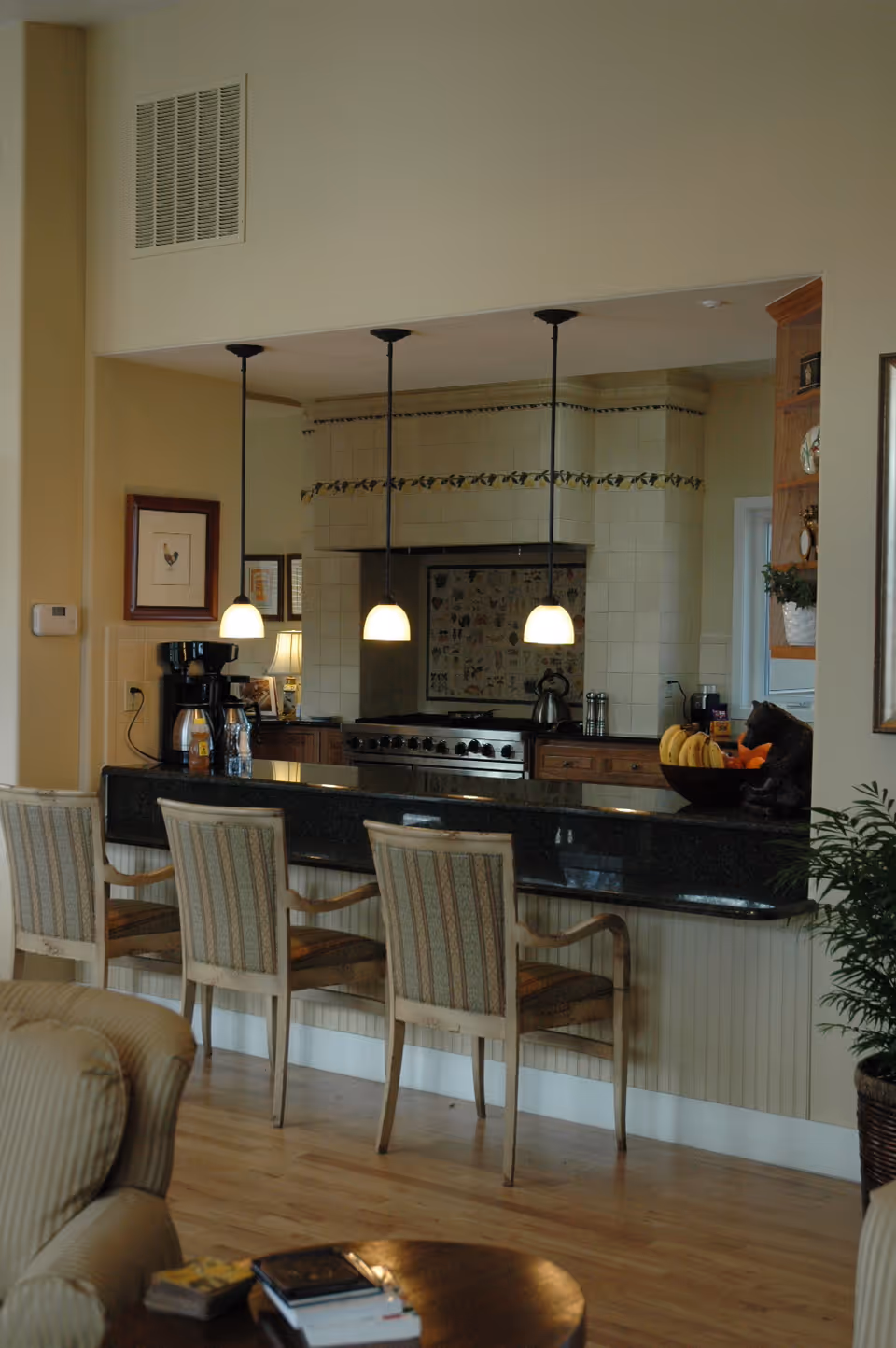 Interior view of a kitchen area with a black granite countertop bar and three wooden chairs with patterned cushions. Above the counter, three pendant lights hang from the ceiling. The kitchen features a large stove with a tiled backsplash and decorative border. A coffee maker and a bowl of bananas are visible on the counter. The foreground includes part of a beige armchair and a round wooden table with books on it.