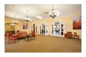 A spacious living room area with beige carpet and yellow walls. The room features multiple seating options including red and brown upholstered chairs and sofas arranged around small tables. There are chandeliers hanging from the ceiling and large arched windows with glass panes along one wall. Artwork is displayed on the walls, and the room has a warm, inviting atmosphere.
