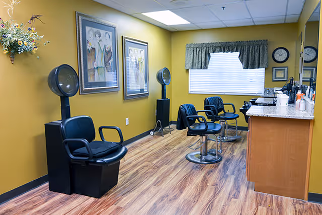 Interior of a hair salon with three black salon chairs, two hair dryers, framed artwork on mustard yellow walls, a window with green valance, and a countertop with hair care products and mirrors.