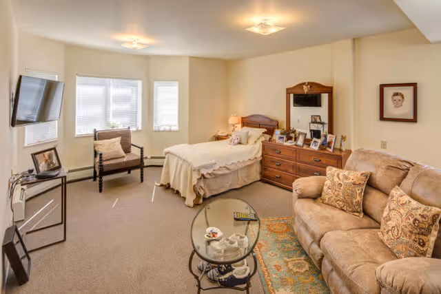 A cozy assisted living facility room with a single bed covered in a beige blanket, a wooden dresser with a mirror and framed photos, a beige sofa with patterned cushions, a glass coffee table with a remote and cups, a wooden chair near a bay window with blinds, and a wall-mounted flat-screen TV.