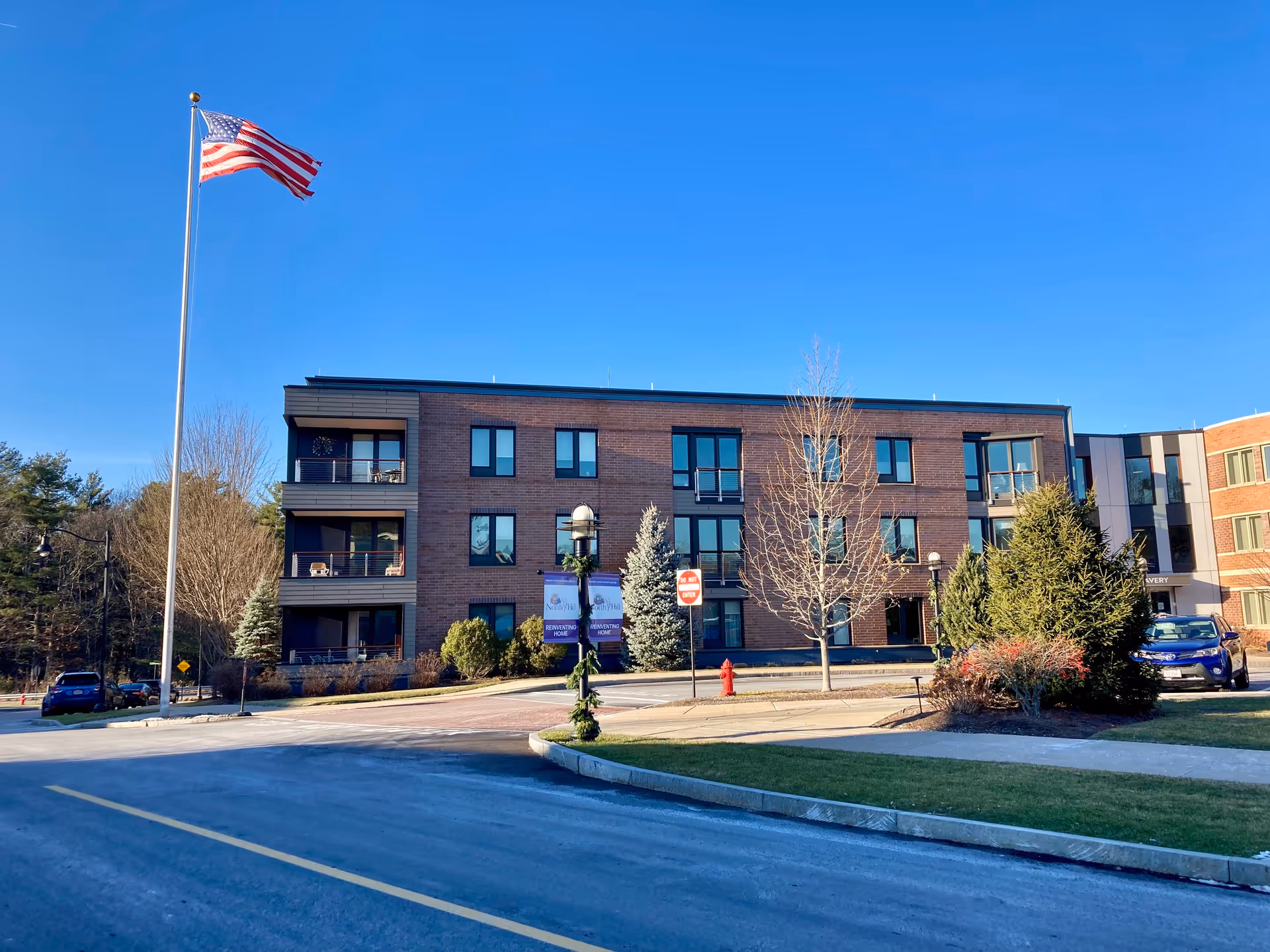 Exterior view of a three-story brick building at North Hill Retirement Community with multiple windows and balconies. An American flag is flying on a tall flagpole in front of the building. There are trees, shrubs, and a small parking area with cars visible. The sky is clear and blue.