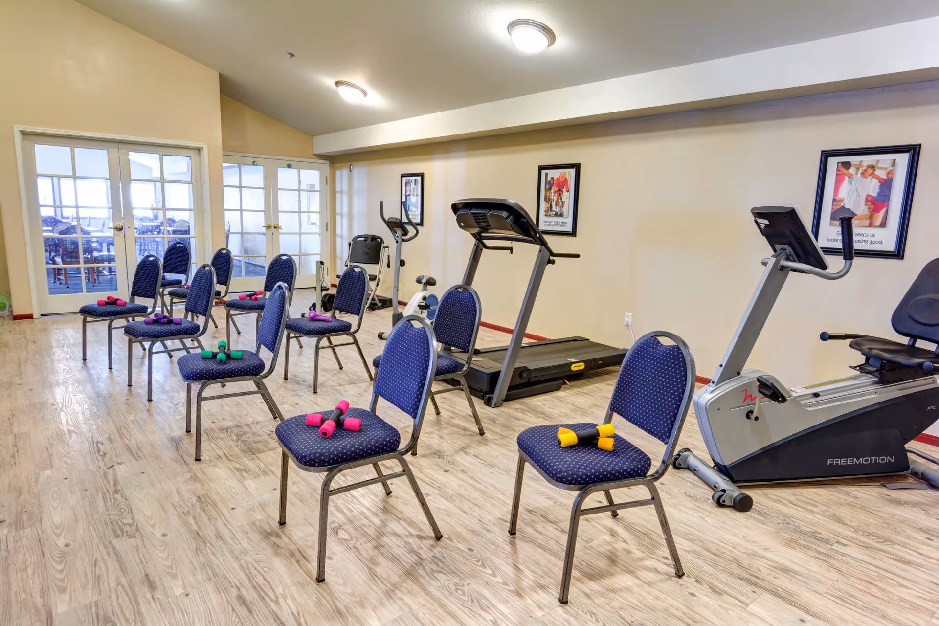 Exercise room with several blue chairs arranged in rows, each chair holding pairs of colorful dumbbells. The room has wooden flooring, two treadmills, and an exercise bike. There are framed motivational posters on the beige walls and glass double doors leading to another room.