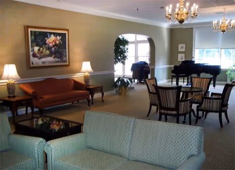 Spacious senior living common room with sofas, armchairs, a dining table, chandeliers, and a piano.