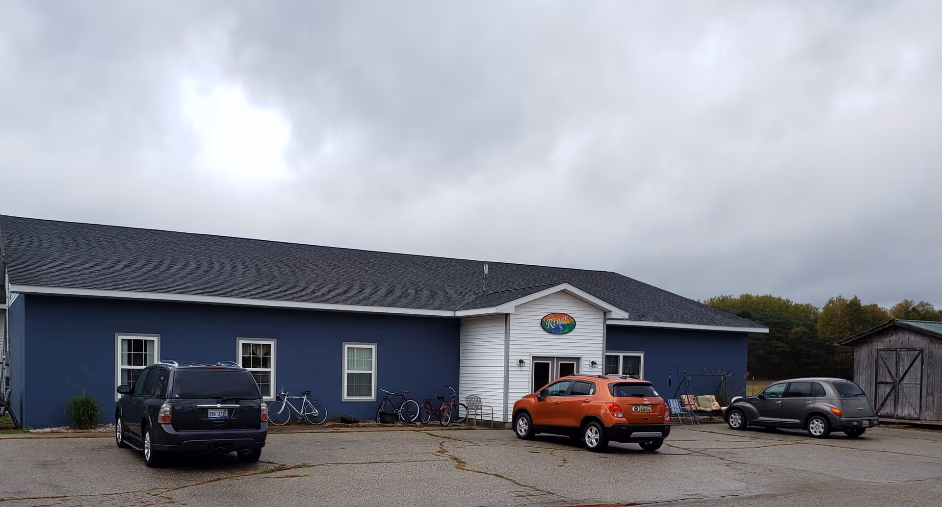 Exterior view of a single-story building with blue walls and a white entrance section under a cloudy sky. Three cars are parked in front of the building, and several bicycles are lined up against the wall near the entrance. There is a wooden shed to the right and some greenery in the background. A colorful oval sign above the entrance reads 'The River AFC'.