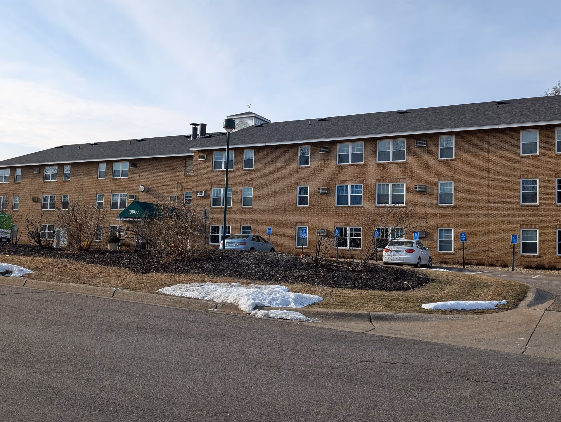 Exterior view of a three-story brick building with multiple windows and a green awning over the entrance displaying the number 10000. There are several handicap parking spaces with cars parked in front. Some patches of snow are visible on the grass near the curb.