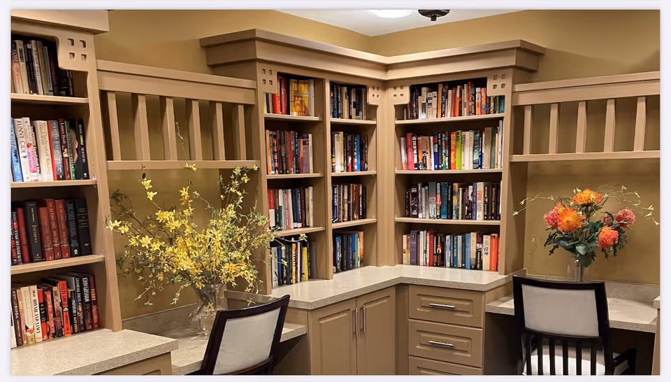 Corner built-in library with beige shelves, books, and two chairs in a senior living facility.