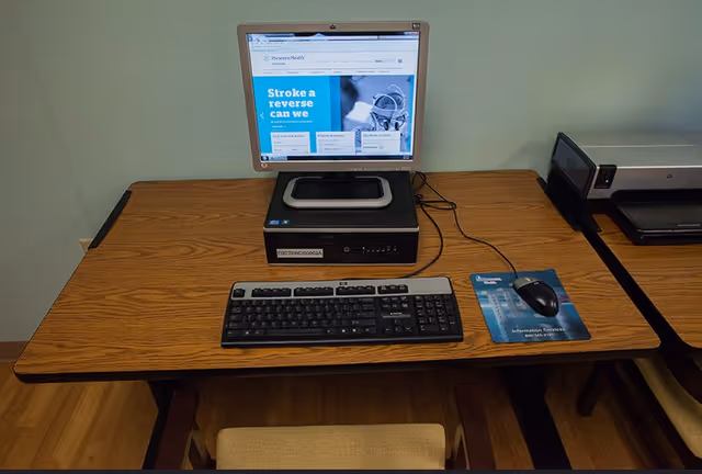 A desktop computer setup on a wooden table with a keyboard, mouse on a blue mouse pad, and a monitor displaying a health-related website. A printer is visible on the adjacent table.