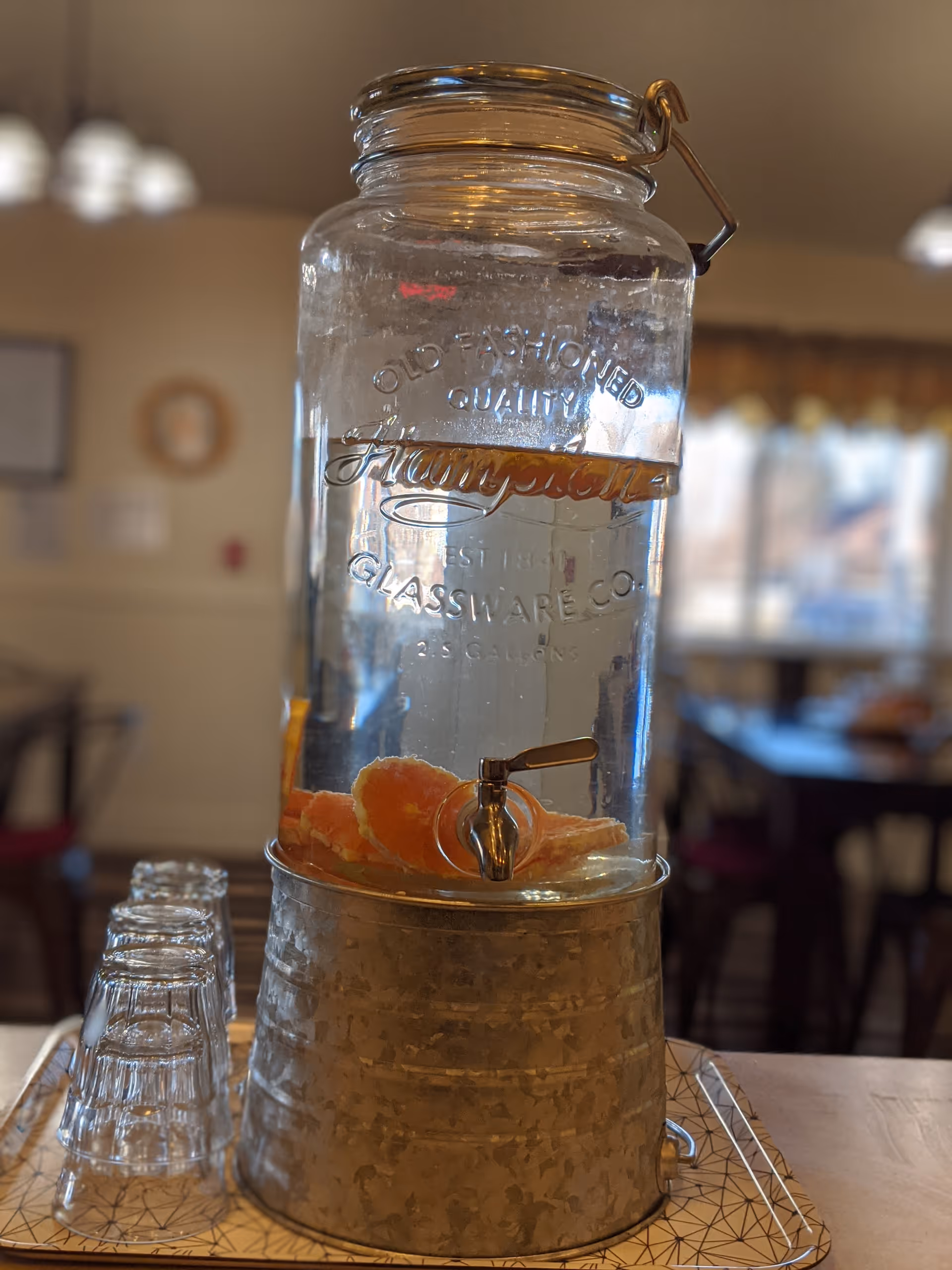 A large glass beverage dispenser filled with water and slices of orange, placed on a metal stand on a tray. Several upside-down drinking glasses are arranged next to the dispenser on the tray. The background shows a softly focused dining area with tables, chairs, and windows.