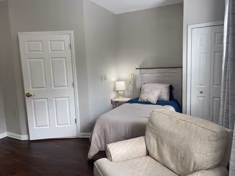 A small bedroom with a single bed against the wall, featuring a gray headboard, blue and gray bedding, and a pillow. Next to the bed is a white nightstand with a lamp and a small decorative item. In the foreground, there is a beige armchair. The room has hardwood floors, light gray walls, a closed white door, and a closet with white double doors. A window with gray curtains is partially visible on the right side.