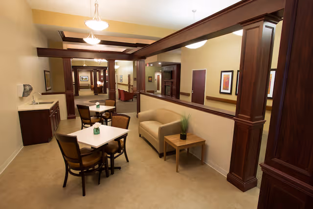 Warmly lit communal dining and seating area with small tables and chairs, a loveseat and dark wood columns.