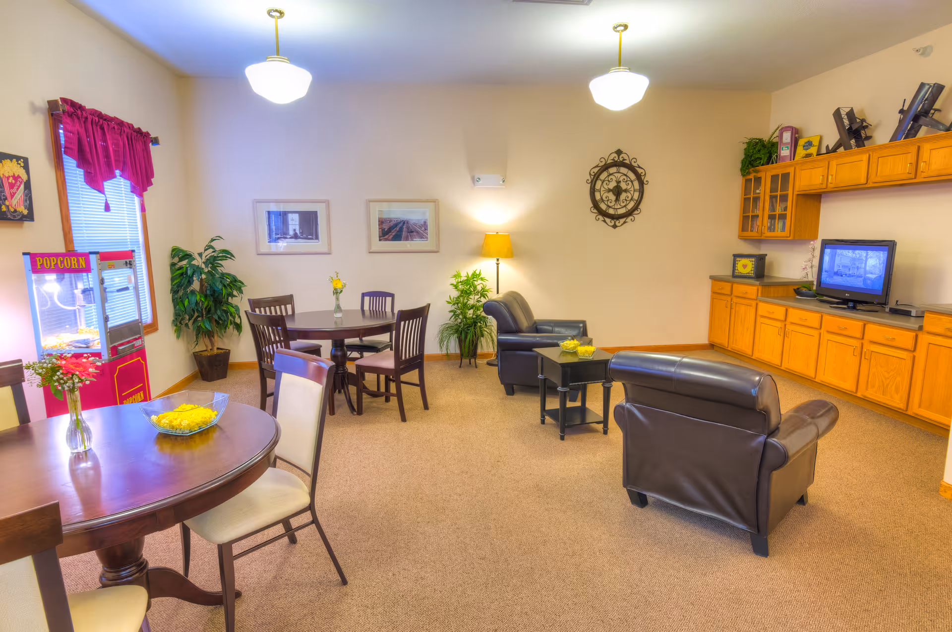 A cozy common area with two black leather armchairs facing a small table with a bowl of popcorn. There is a TV on a wooden cabinet along the right wall. Two round tables with chairs are positioned on the left side, one with a vase of flowers and a bowl of popcorn. A popcorn machine is near the window with red curtains. The room is decorated with plants, framed pictures, a wall clock, and a floor lamp.