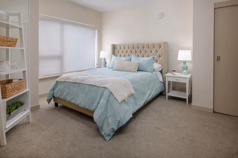A neatly made bed with a tufted beige headboard, light blue bedding, and decorative pillows in a carpeted bedroom. There are two bedside tables with matching blue lamps and a white shelving unit with baskets and a small plant near a window with a closed blind.