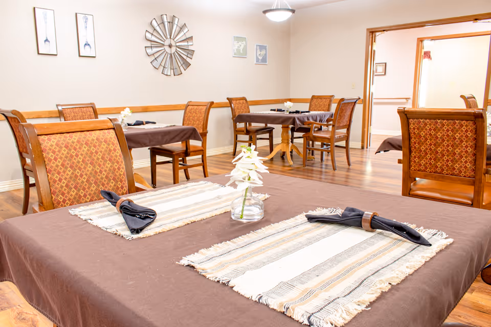 Dining room with several tables and wooden chairs set with placemats, napkins, and small flower centerpieces.