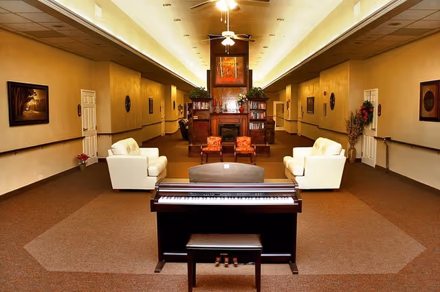 A spacious common area in a senior living facility featuring a digital piano with a bench in the foreground, two white armchairs facing each other, two red patterned chairs, a wooden fireplace with bookshelves on either side, and warm lighting from ceiling fixtures. The walls are decorated with framed artwork and plants.
