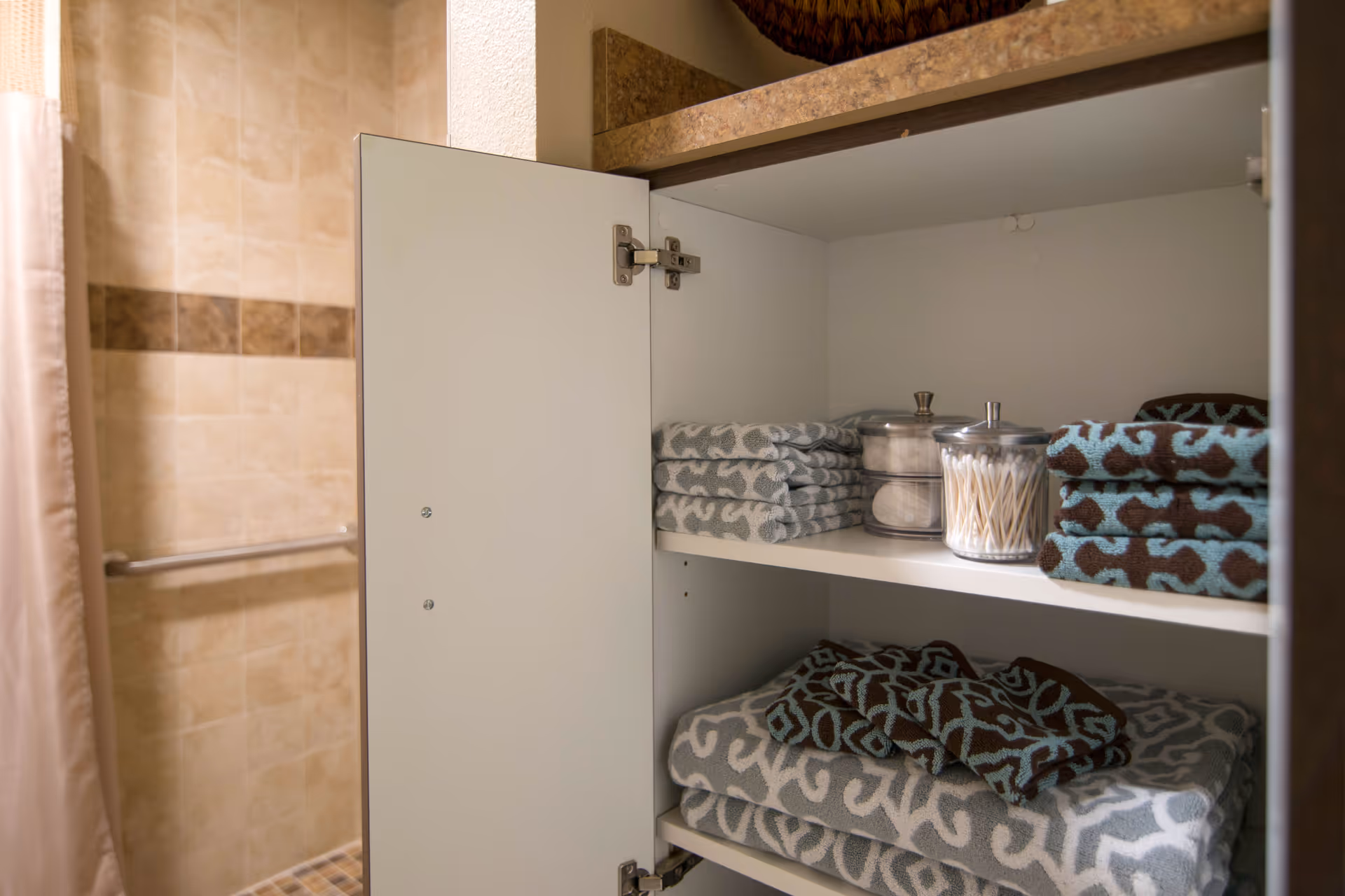 Open bathroom cabinet with neatly folded patterned towels and two glass containers, one holding cotton balls and the other holding cotton swabs. In the background, a tiled shower area with a grab bar is partially visible.
