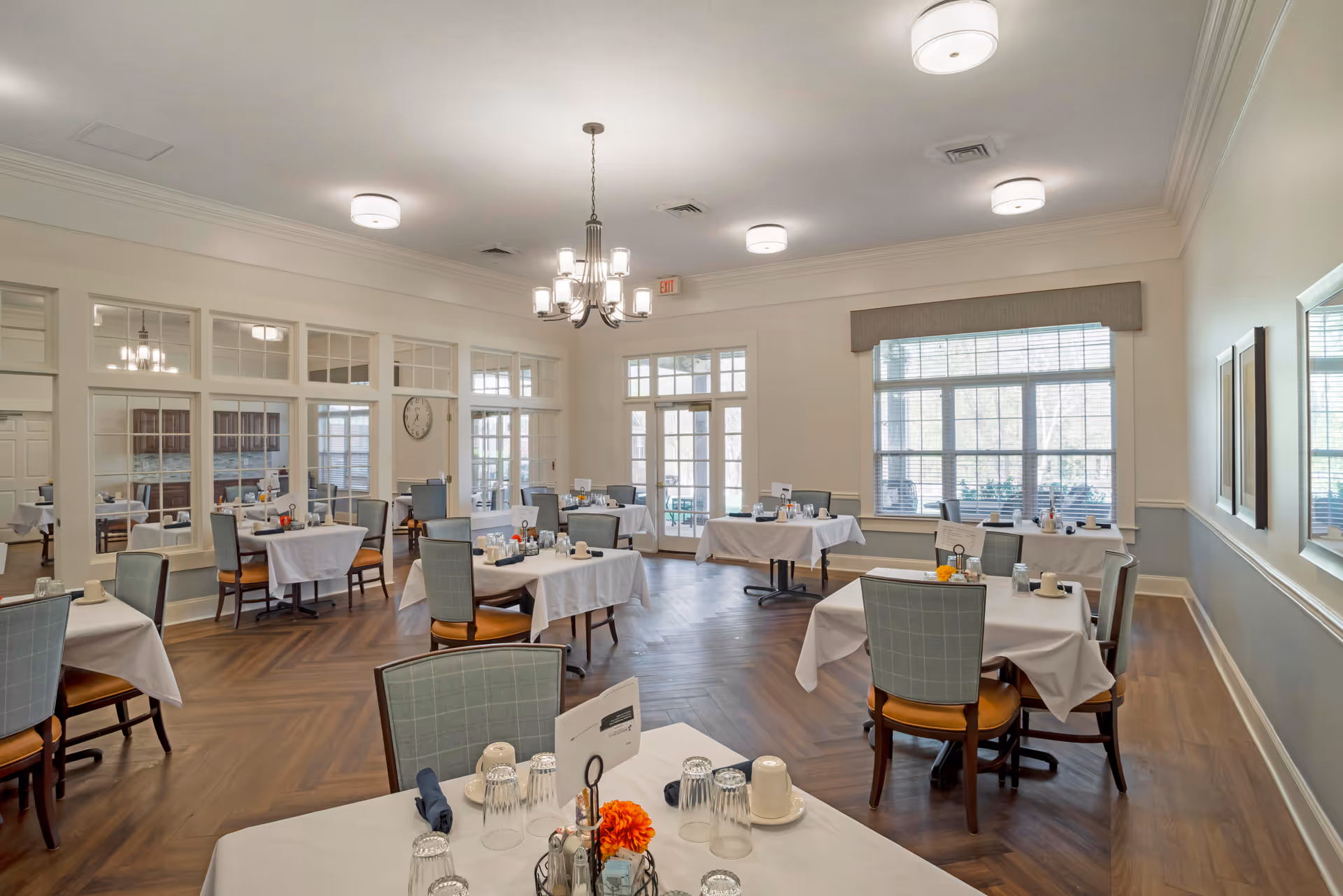 A bright and spacious dining room with multiple tables covered in white tablecloths, each set with glasses, cups, and condiments. The room features large windows allowing natural light to fill the space, a chandelier hanging from the ceiling, and wooden flooring with a herringbone pattern. Chairs with light green upholstery and wooden legs surround the tables.