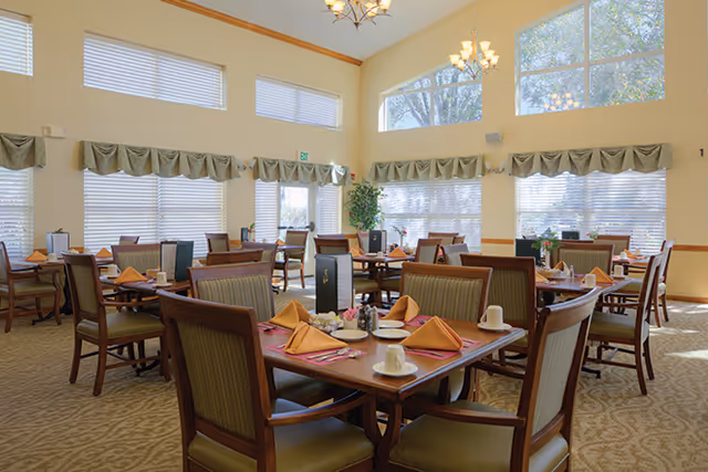 Bright dining room with multiple tables set with napkins and place settings, high windows, and chandeliers.