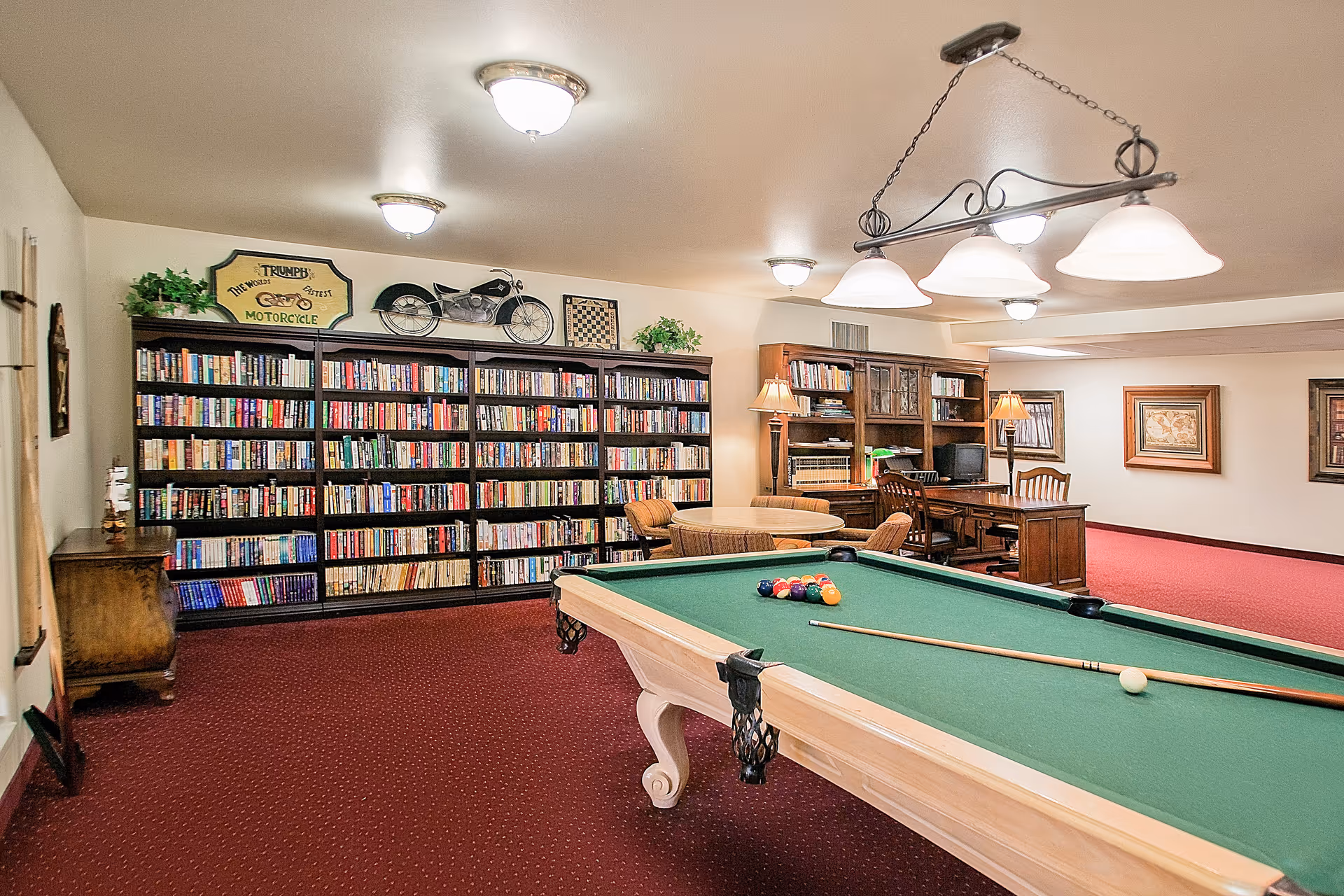 A recreational room with a pool table in the foreground, a large bookshelf filled with books along the back wall, a round table with chairs, and a wooden desk with a computer and lamp. The room has red carpet, white walls, and ceiling lights.