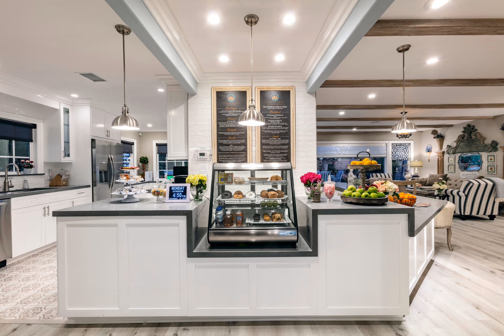 A bright and modern kitchen area with white cabinets, gray countertops, and stainless steel appliances. In the center, there is a display case with assorted pastries and muffins, flanked by two pendant lights. The kitchen opens into a living room area with striped and tufted sofas, decorative wall art, and a large window showing an outdoor patio. Various flowers and fruit bowls decorate the countertops.
