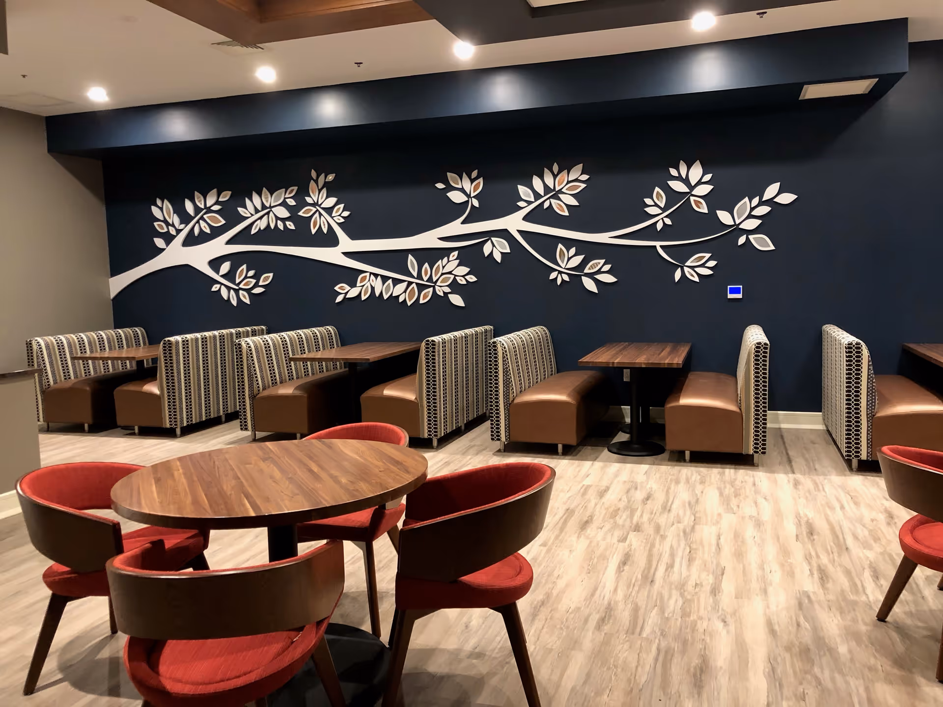 Interior dining area with wooden tables and a mix of red cushioned chairs and patterned booth seating. The back wall is dark blue with a large decorative white tree branch with leaves mounted on it. The floor has light wood-style flooring and the ceiling has recessed lighting.