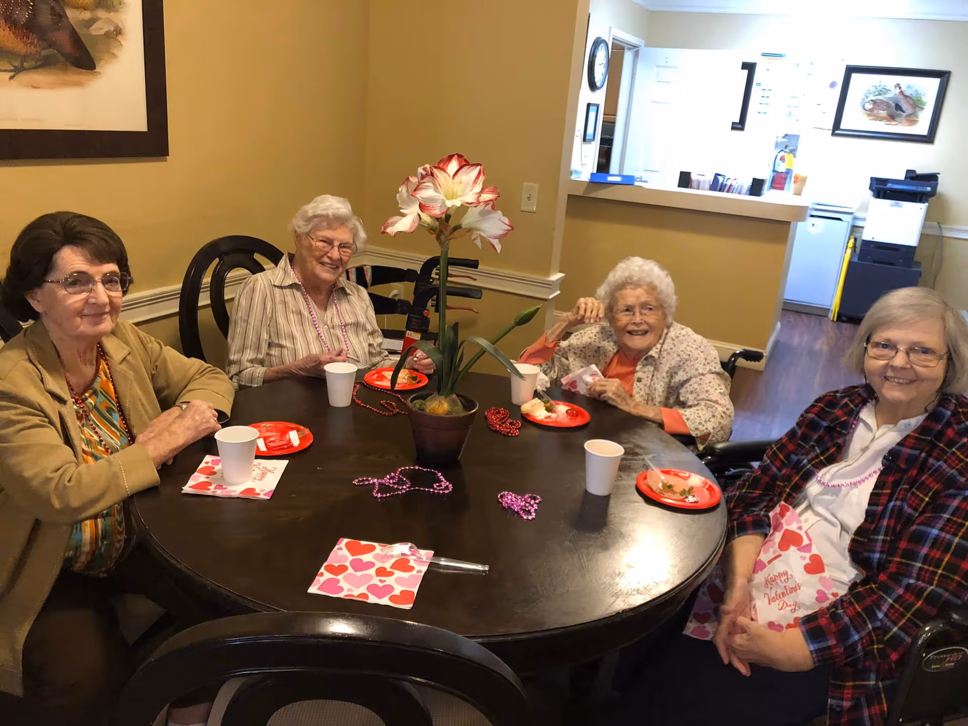 Four elderly women sit around a dining table with heart-themed napkins and plates and a potted flower centerpiece.
