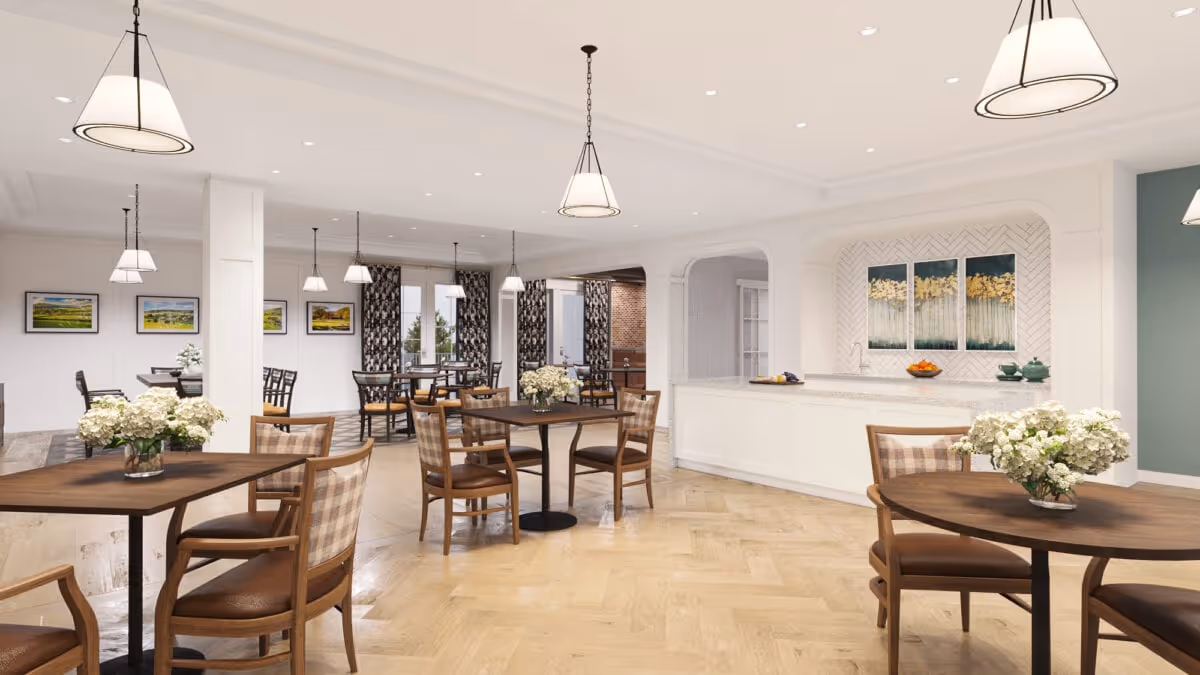 A bright and spacious dining room with multiple wooden tables and chairs arranged neatly. Each table has a vase with white flowers. The room features light wood flooring, white walls, and several pendant lights hanging from the ceiling. There are large windows with patterned curtains allowing natural light to enter. On one side, there is a white counter with decorative artwork above it.