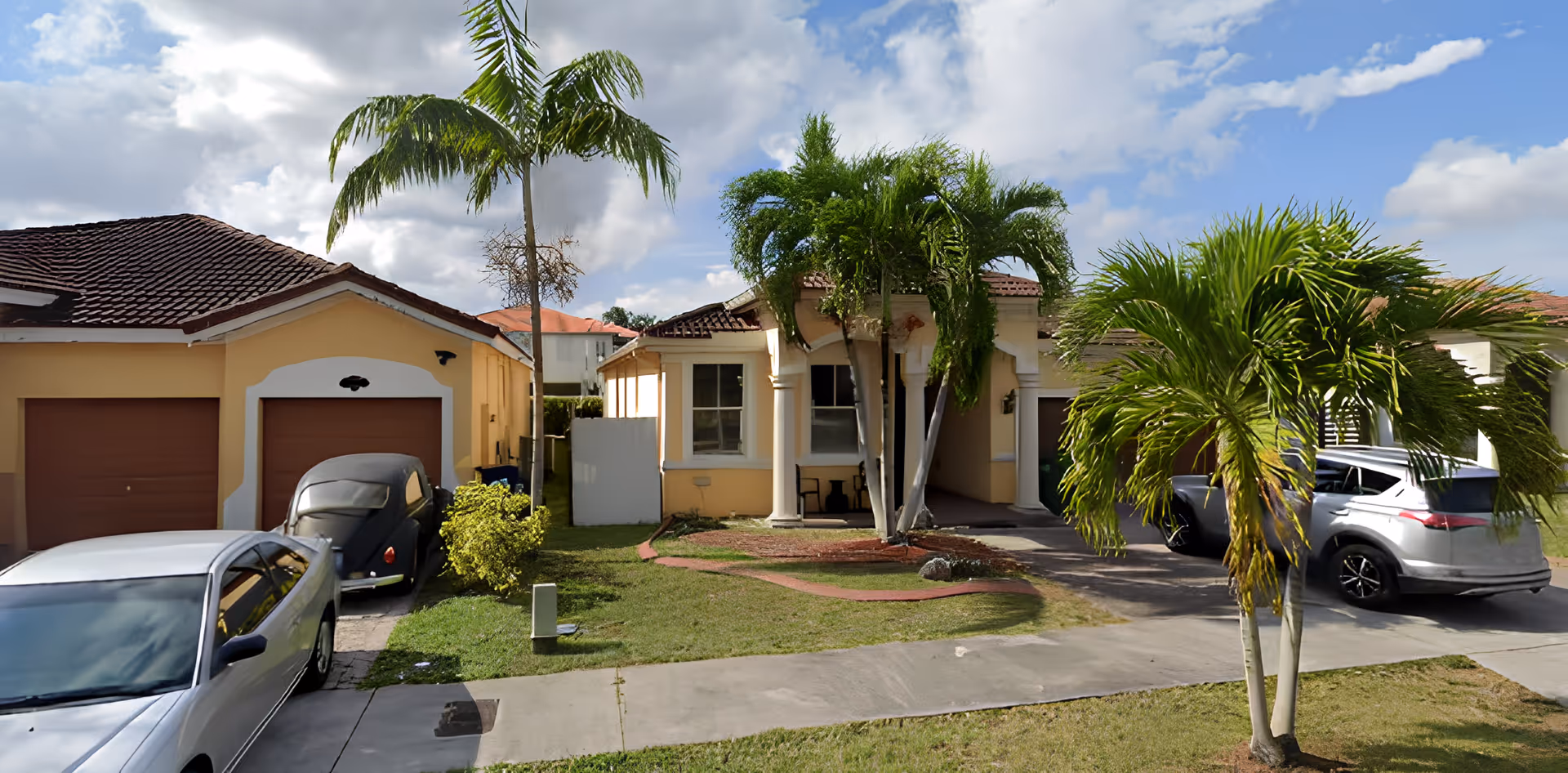 Front exterior view of a single-story residential building with a tiled roof, two palm trees in the front yard, a driveway with parked cars, and a small lawn area under a partly cloudy sky.