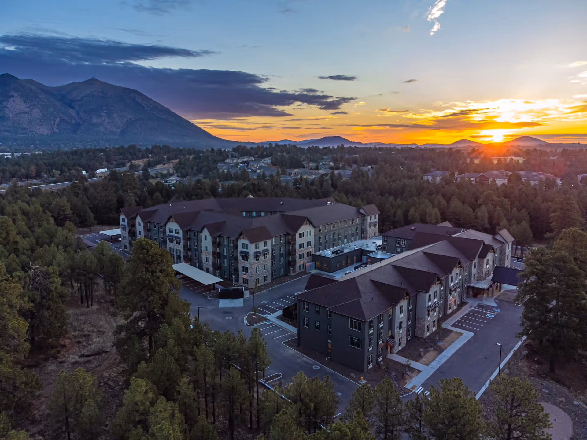 Aerial view of The Bluffs of Flagstaff Senior Living facility at sunset, surrounded by trees and mountains in the background with a colorful sky.