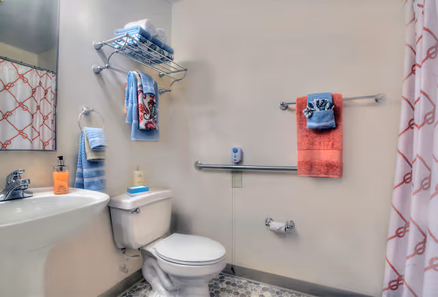 Accessible bathroom with a pedestal sink, toilet, grab bar, towel racks with folded towels, and a patterned shower curtain.