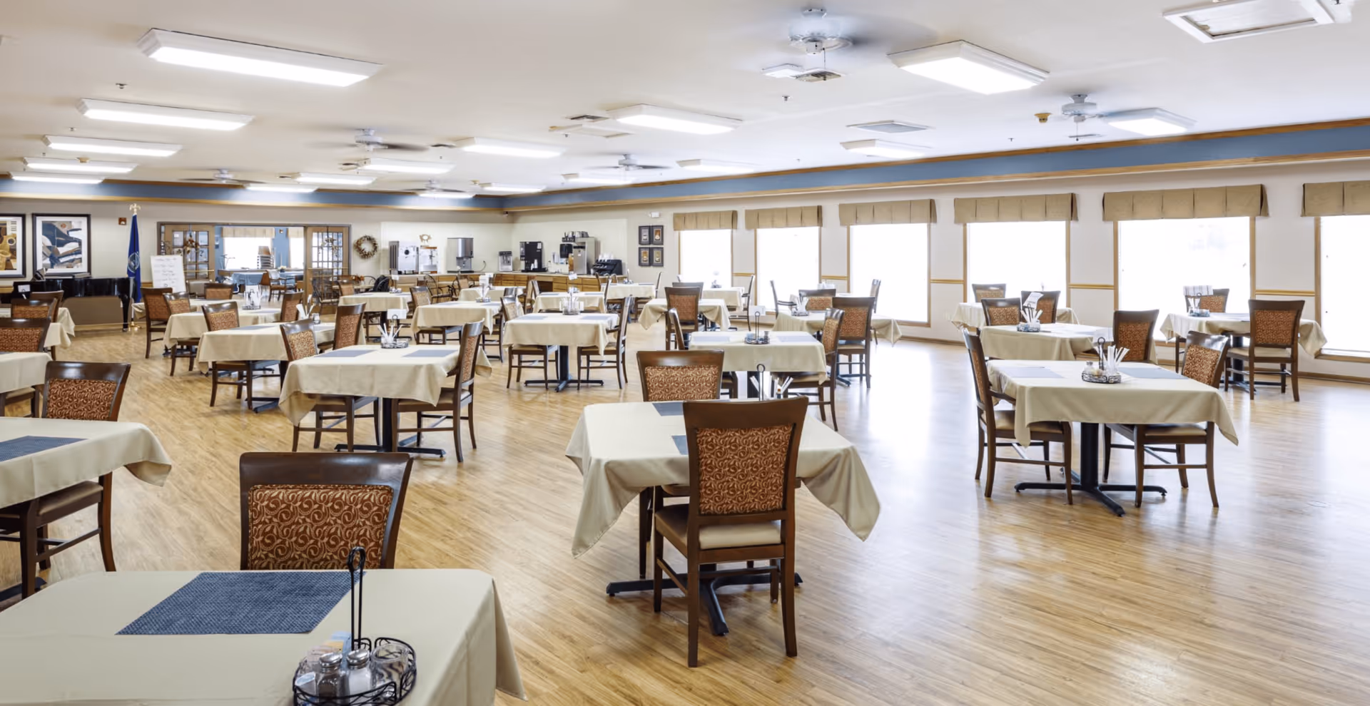 A spacious dining room with multiple tables covered in beige tablecloths and set with placemats, salt and pepper shakers, and napkin holders. The room has wooden floors, large windows with beige valances, and bright overhead lighting. There is a coffee and beverage station along the far wall and framed artwork on the walls.