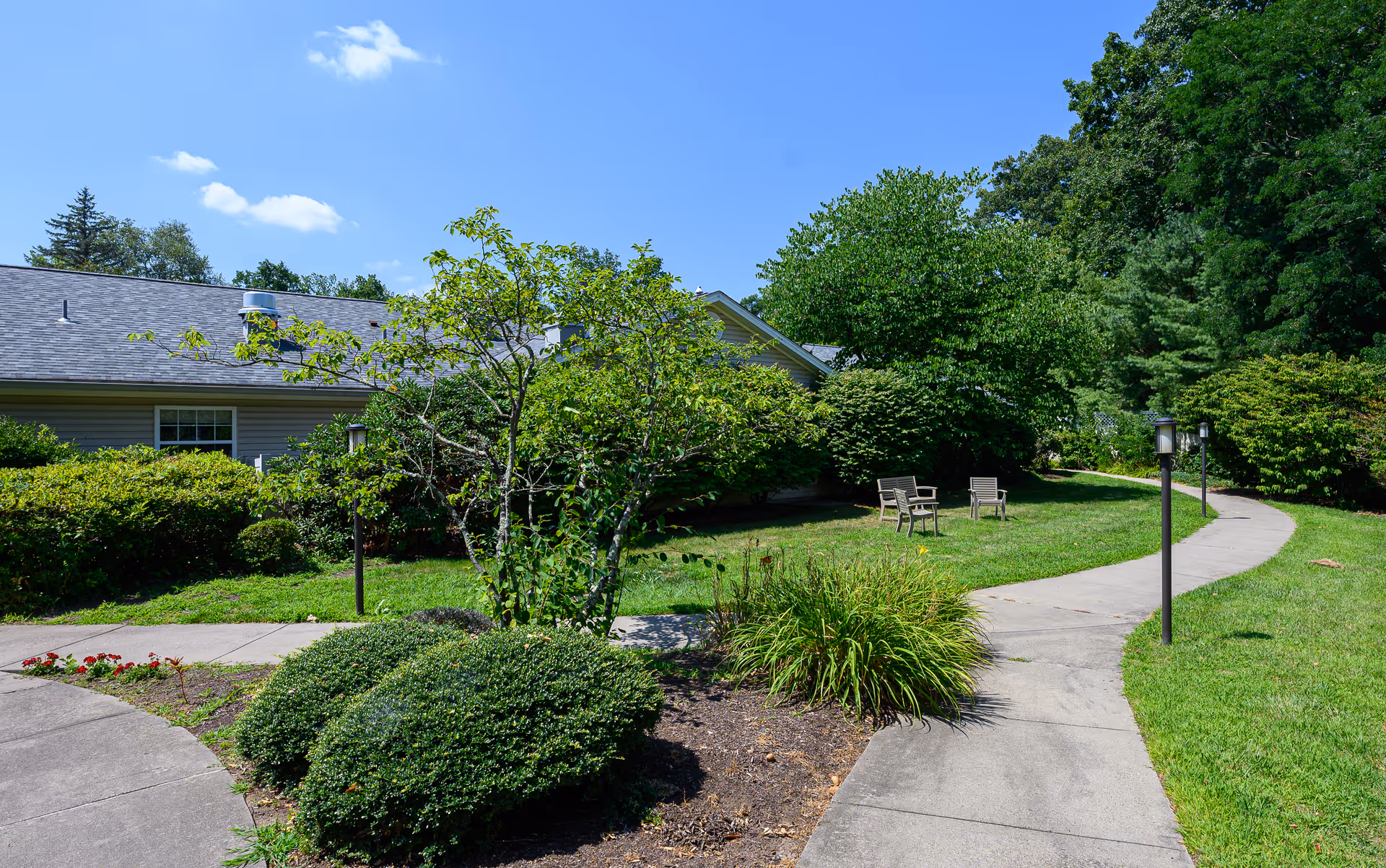 A sunny outdoor garden area with a curved concrete pathway, green bushes, small trees, and two wooden chairs on a grassy lawn. The side of a building with a gray roof and beige siding is visible in the background under a blue sky with a few clouds.