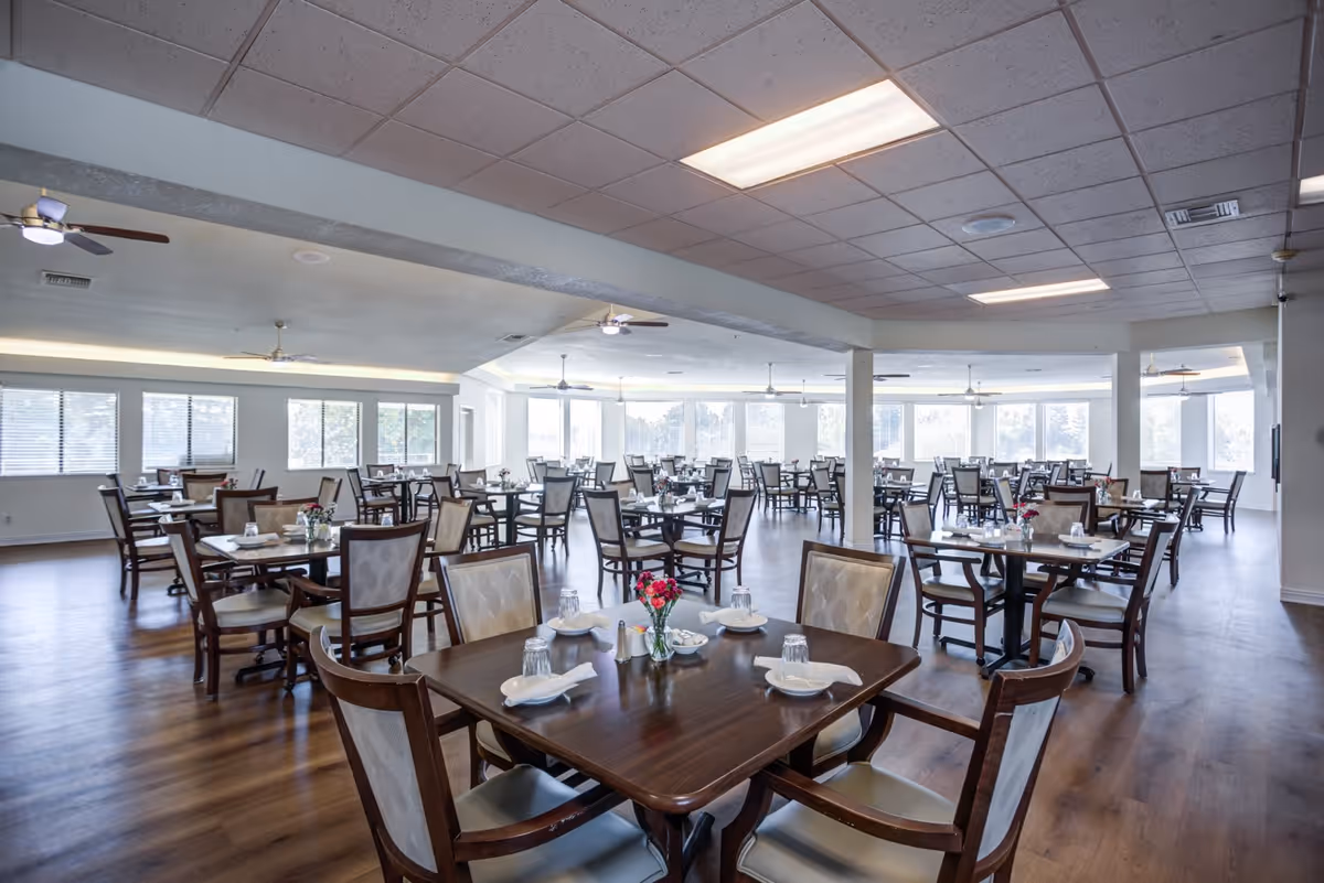 A spacious dining room with multiple wooden tables and chairs arranged neatly. Each table is set with white napkins, glasses, and small flower vases. Large windows allow natural light to fill the room, and ceiling fans are mounted on the ceiling.