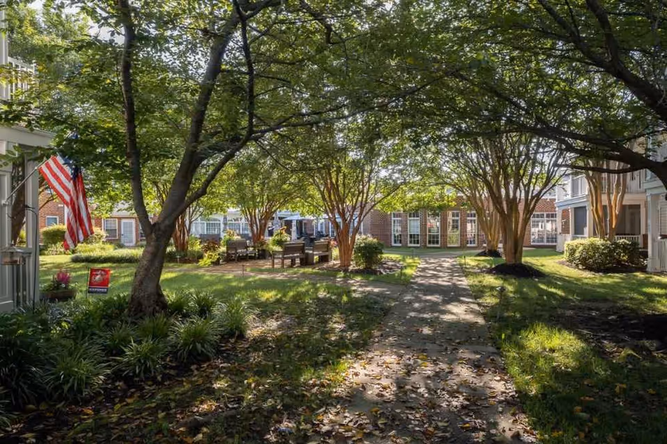 A peaceful outdoor courtyard area at Chancellor's Village with a paved walkway, green grass, several trees providing shade, benches arranged in a seating area, and buildings with large windows surrounding the space. An American flag is displayed on the left side near the entrance.