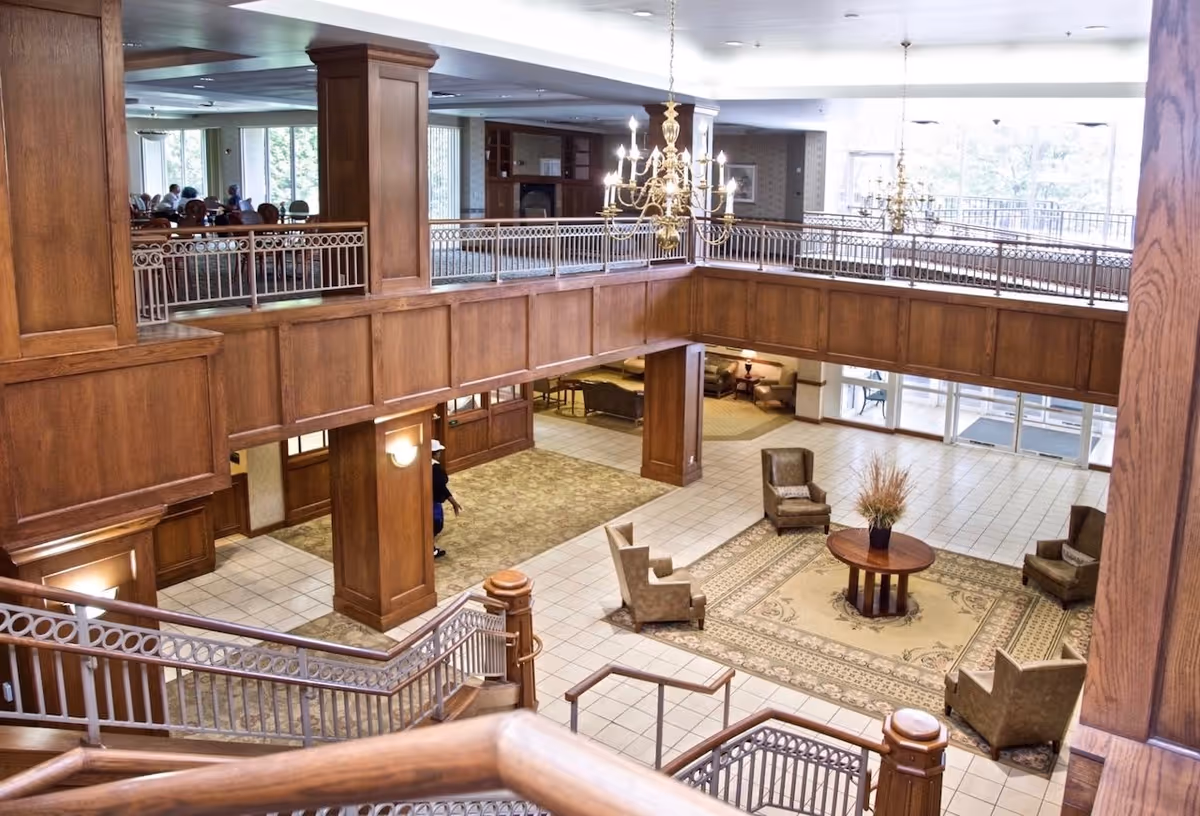 View of a spacious senior living facility lobby with wooden paneling, a large chandelier, and seating areas with armchairs arranged around a round table with a plant centerpiece. There is a staircase leading down to the tiled floor area and a dining area visible on the upper level with people seated at tables.