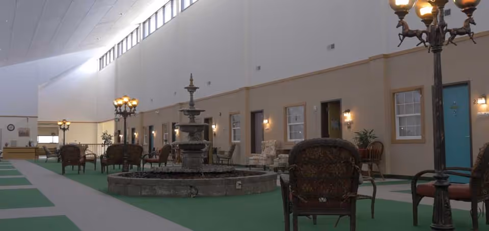 Indoor common area with green carpet and a central stone fountain surrounded by chairs and vintage-style lamp posts. The walls have multiple doors and windows, and the ceiling has high windows letting in natural light.