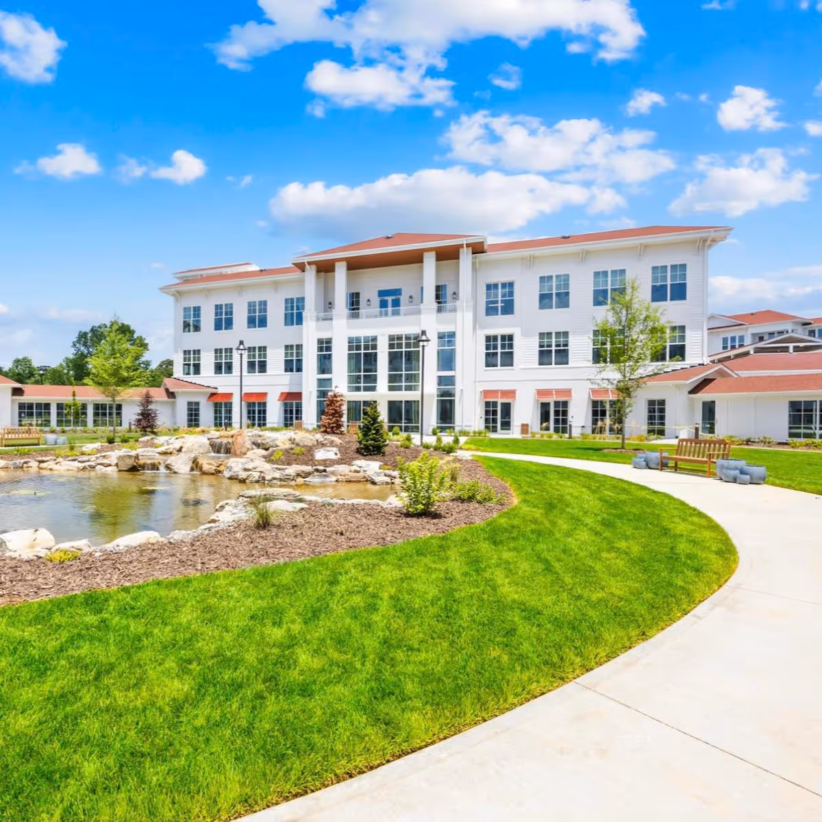 Three-story white senior living building with a landscaped pond, green lawn and curved walkway under a blue sky.