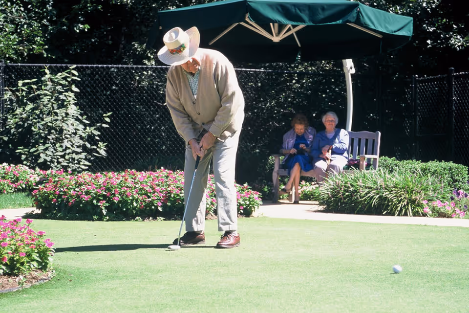 An elderly man wearing a wide-brimmed hat and beige cardigan is playing putting golf on a green lawn surrounded by flower beds. In the background, two elderly women are sitting on a bench under a large green umbrella, with bushes and a black chain-link fence behind them.