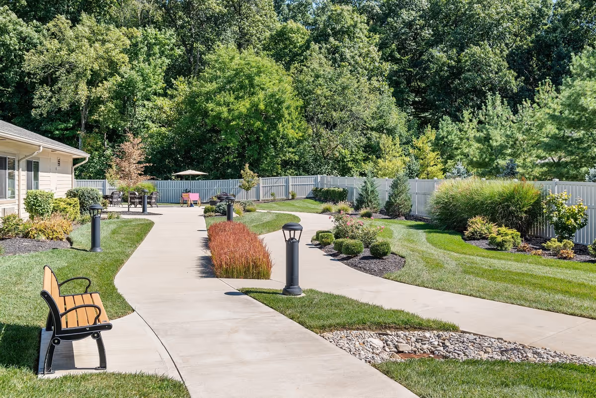 A well-maintained outdoor garden area with a winding concrete pathway, green grass, various shrubs and plants, a wooden bench on the left side, and a table with chairs and an umbrella in the background. The area is enclosed by a white fence and surrounded by tall trees.