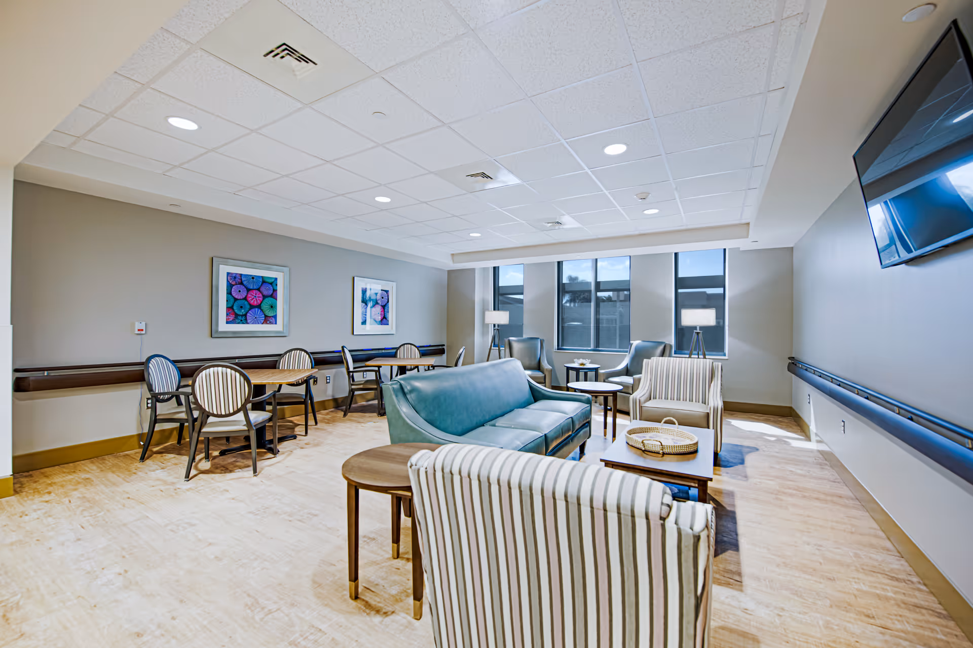 A bright and spacious common area in a senior living facility featuring a teal sofa, striped armchairs, wooden tables, and chairs along the wall. The room has large windows allowing natural light, two framed colorful artworks on the wall, and a flat-screen TV mounted on the right wall.