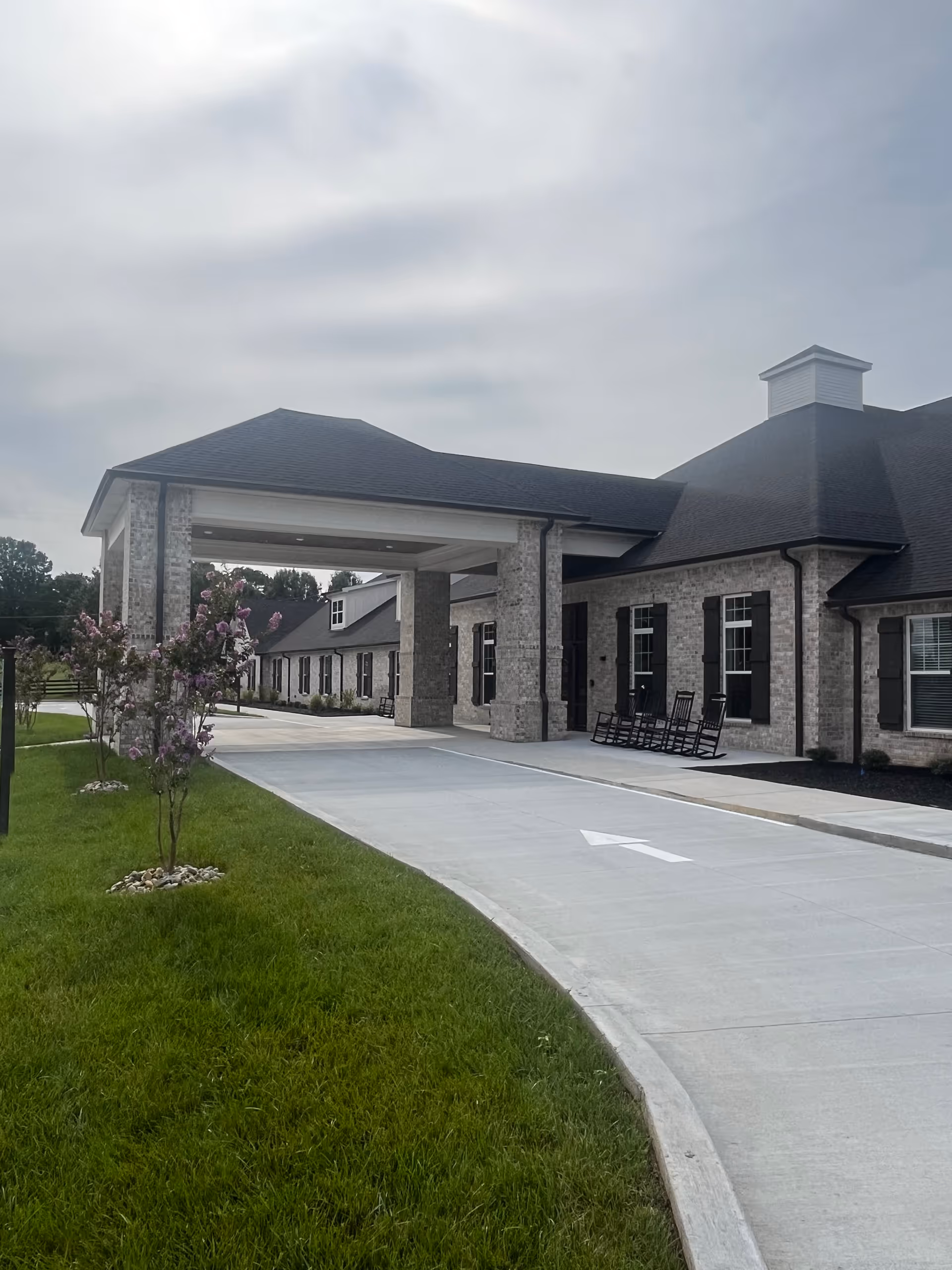 Exterior view of The Pearl at Fairview facility showing a covered entrance with a driveway, green lawn with small flowering trees, and a row of rocking chairs on the porch of a brick building under a cloudy sky.