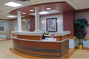 Reception area inside Meadow Woods Nursing And Rehab Center featuring a curved wooden desk with white countertop, a computer, a chair, decorative columns, framed artwork on the wall, and a potted plant in the corner.