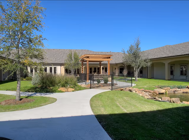 Outdoor courtyard area at Volante Senior Living of Grapevine featuring a paved walkway, green grass, trees, shrubs, a small pond with rocks, and a wooden pergola structure near the building under a clear blue sky.