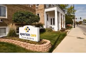 Exterior view of a brick building with a white entrance porch and a landscaped area featuring a sign that reads 'aperion care PLUM GROVE' next to a sidewalk and street.