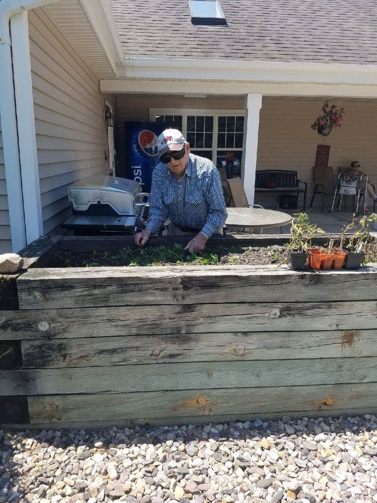 An elderly man wearing a checkered shirt, sunglasses, and a white cap is tending to plants in a raised wooden garden bed outside a building. Behind him, there is a patio area with chairs, a table, a grill, and a Pepsi vending machine. The ground is covered with small rocks.
