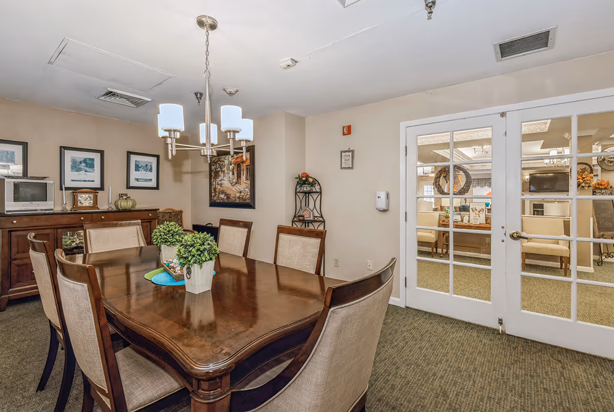 A well-lit dining room with a large wooden table and upholstered chairs, a chandelier overhead, and French doors opening to a lounge area.